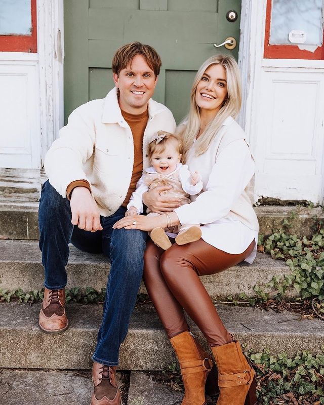 Lauren Wirkus in a white shirt, brown leather pants and brown boots posing with her husband and daughter in white jackets.